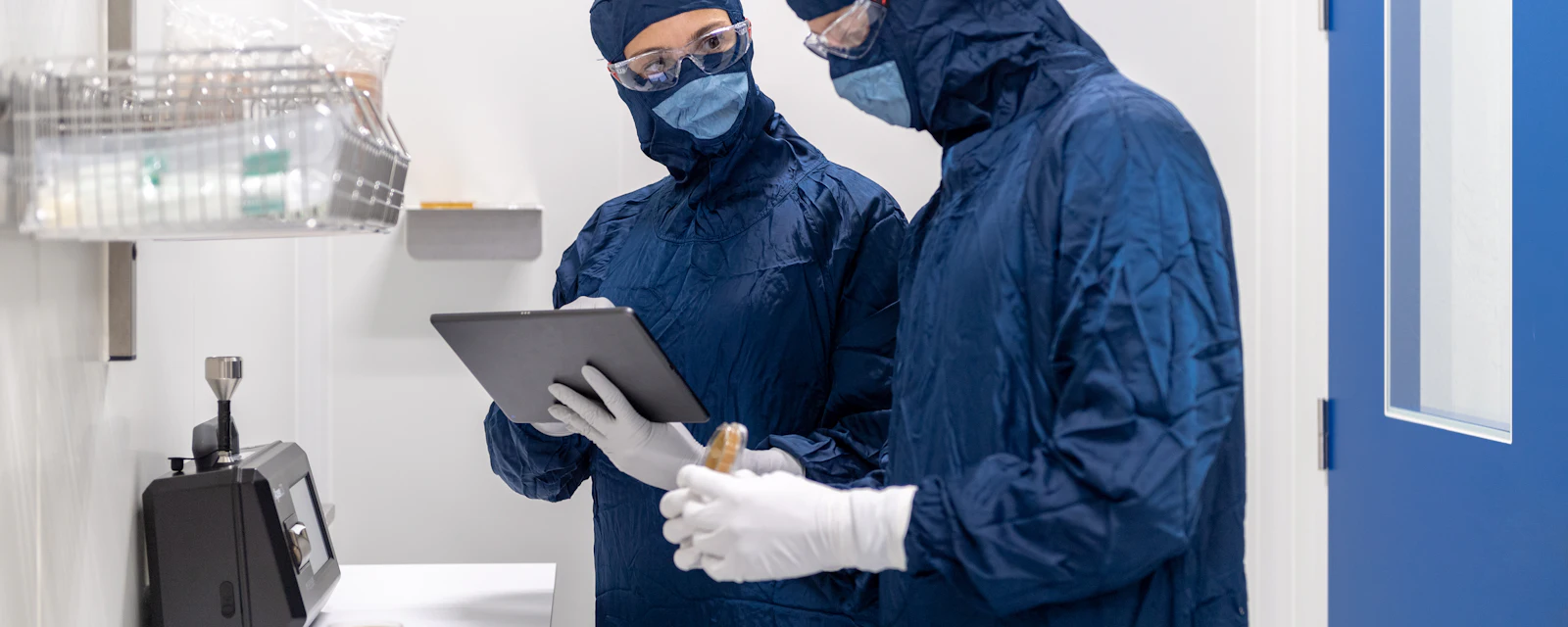Two laboratory technicians in cleanroom suits reviewing data on a tablet